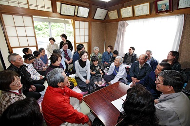 Mrs. Enomoto in a small room surrounded by around 20 seated men, women and children.