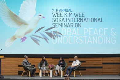 Four people seated on a stage in front of a huge image of a dove with an olive branch.