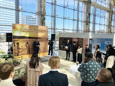 A man stands in front of a large exhibition panel and delivers an address to an audience.