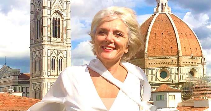 Una mujer en blusa blanca y de pie al aire libre, teniendo de fondo el campanario de Giotto del complejo del Duomo en Florencia. 