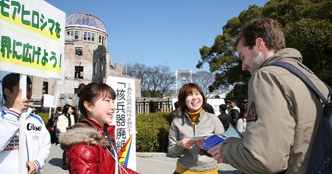 A street scene in which a man prepares to sign a petition presented to him by three young women.