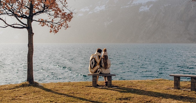 A couple seated on a bench at a lakeside, seen from behind 