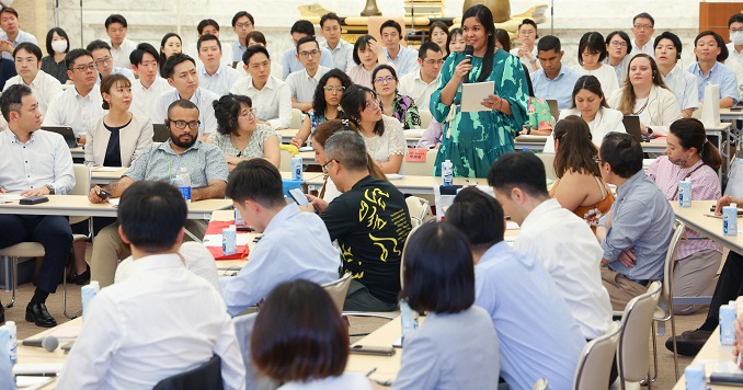 People seated listening to a woman standing and talking.