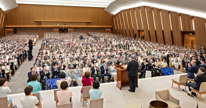 People seated in a large hall facing a speaker at a podium on a stage.