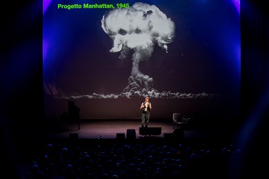 舞台上，1名男士站在有著核爆炸畫面的大螢幕下。