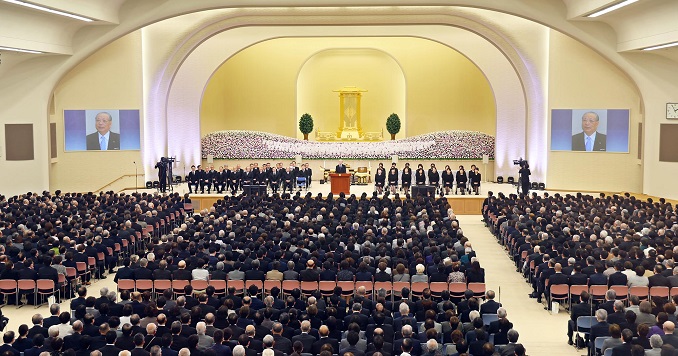People dressed in black in a hall, man giving speech at a podium.