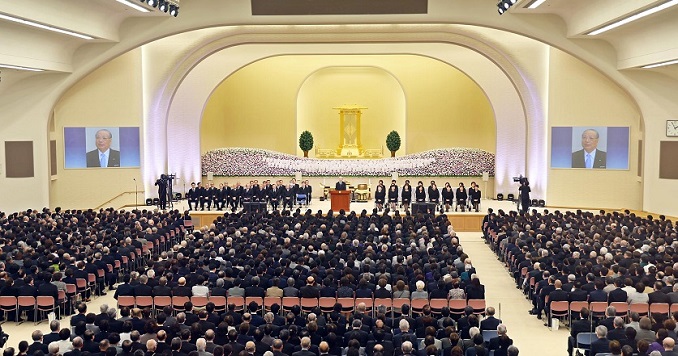 People dressed in black in a hall, man giving speech at a podium.