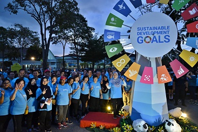 A diverse group of people posing next to a windmill-like structure, the spokes of which display the icons for each of the Sustainable Development Goals.