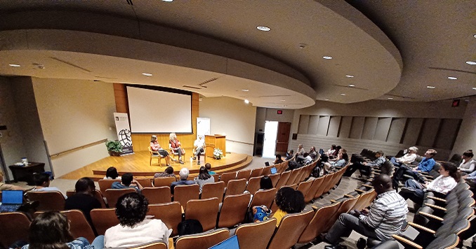 人們坐在會議室理,並且有三名專題演講人坐在台上。
