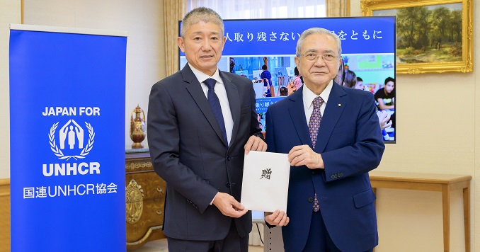兩位男士手拿著信封,站在寫著聯合國難民署的橫幅旁邊。