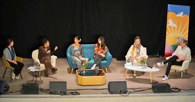 Seis personas sentadas en un escenario, una de ellas sujeta un micrófono y habla por él.