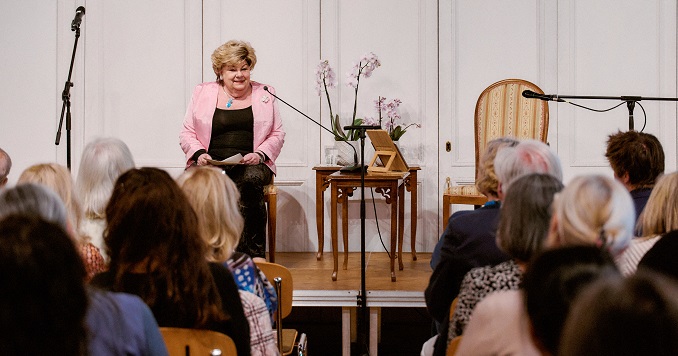 A woman speaking on stage next to some orchids.