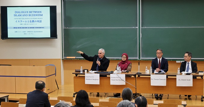 Four people seated at desks facing an audience in a conference room.