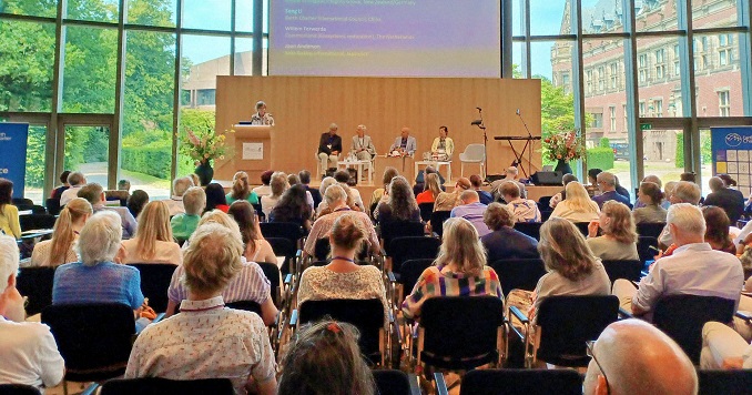 Many people sitting in a conference room facing the stage in a room with large windows.