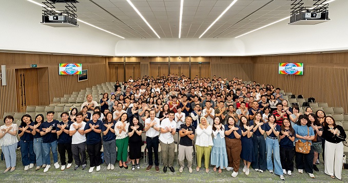 Audience members in an auditorium face the stage, holding their hands up in the shape of a bird, with thumbs linked and fingers spread like wings. 