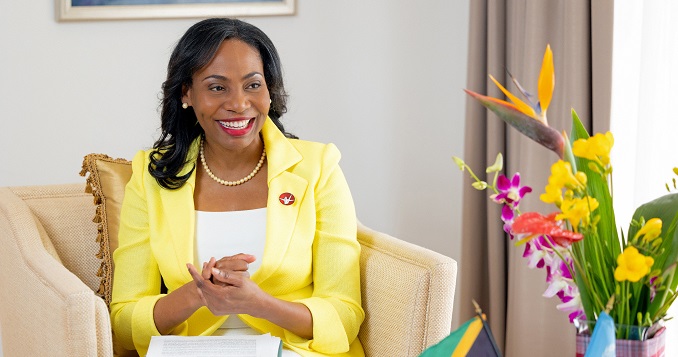 A woman dressed elegantly in a yellow suit, sitting on a chair and smiling.