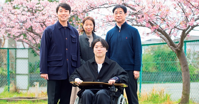 A man in a wheelchair sits with his family under a blooming cherry tree in a park.