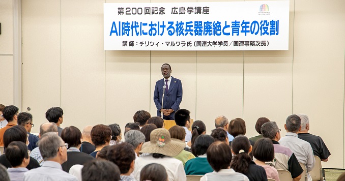 A man in a suit talking into a microphone in front of lots of people sitting down.