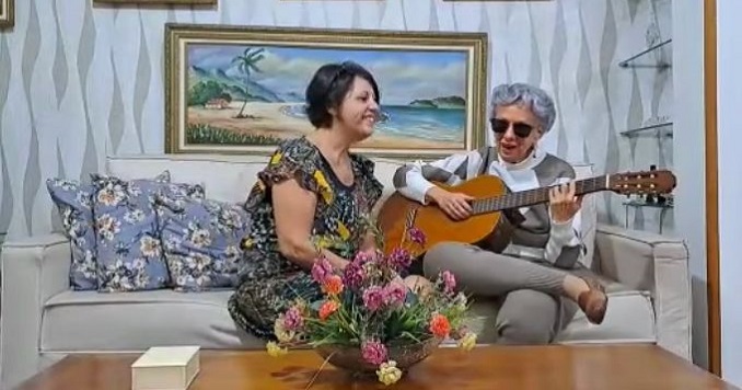A woman with grey hair and dark glasses, playing the guitar for her friend.