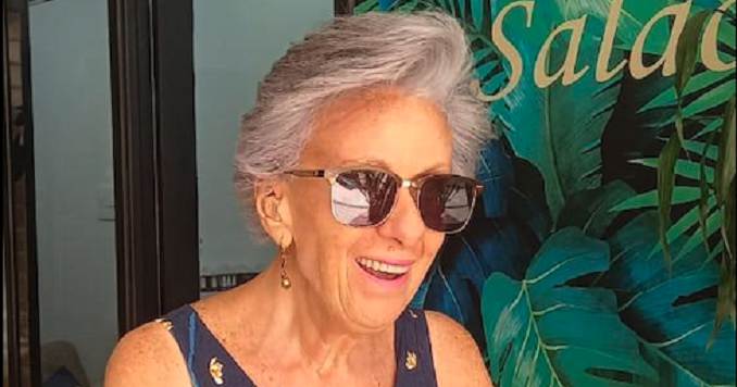 A woman with sunglasses, smiling in front of a painted foliage background.