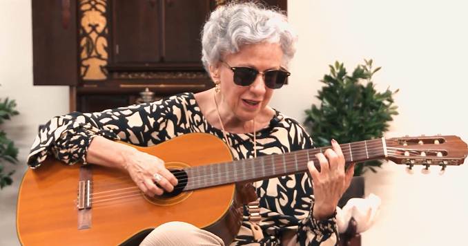 A woman with dark glasses, sitting and playing the guitar.