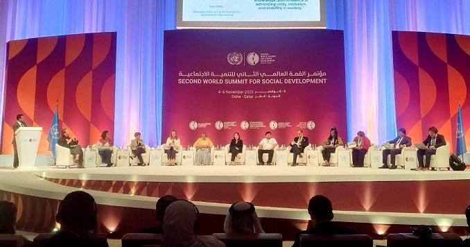 A diverse group of panelists sit on a large stage with several screens behind them.
