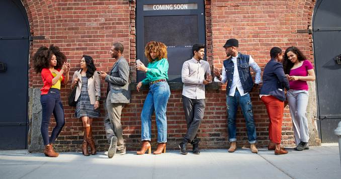 A diverse group of people standing against a brick wall, chatting and checking their phones.