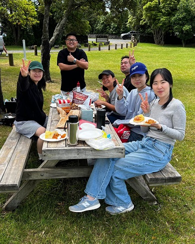 Six people sitting on a bench eating a picnic.
