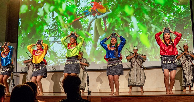 Dancers in vibrant costumes perform on stage before a bright screen depicting a forest and colorful birds.