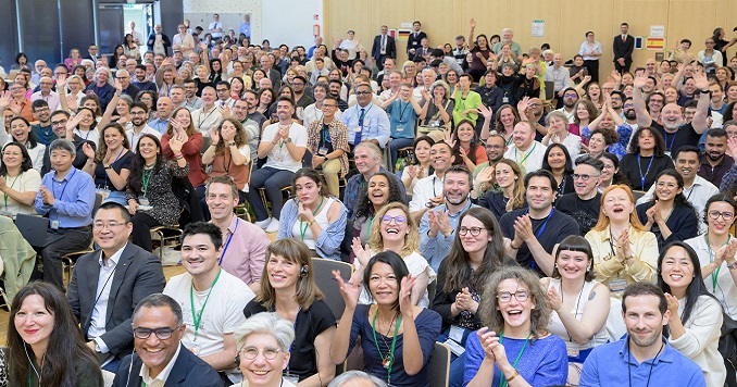 People in a large conference hall smile and wave for a commemorative photo.