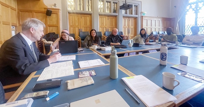 Formally dressed people around a conference table in a room with wood-paneled walls.