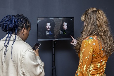 Two people, one looking at her phone and the other at a big screen.