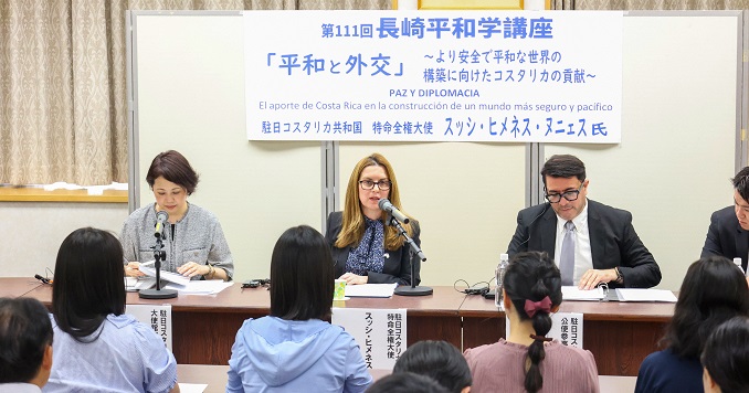 A woman speaks while seated at a table with other panelists, facing an audience.