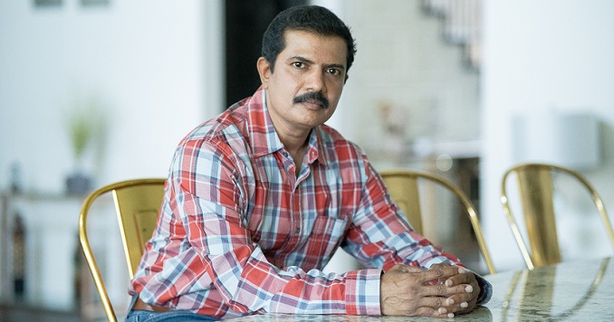 Kiran Malige seated at a table in his home wearing a checked shirt.