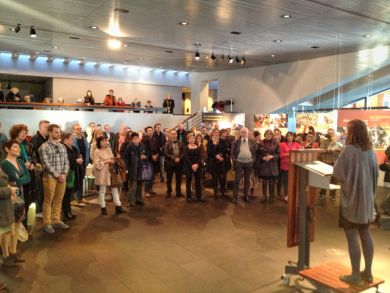 A woman speaks from a podium to a large standing audience in a hall