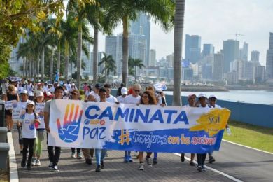 Una columna de personas sonrientes de todas las edades camina por un paseo marítimo bordeado de palmeras con pancartas a favor de la paz