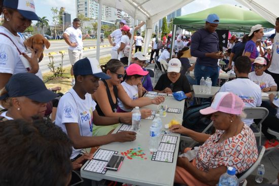 Personas de varias edades se sientan en mesas al aire libre debajo de toldos jugando al bingo y a otros juegos