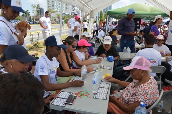 Personas de varias edades se sientan en mesas al aire libre debajo de toldos jugando al bingo y a otros juegos