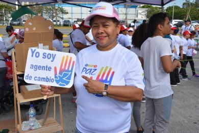 Una mujer mayor posa con un letrero en español que dice “Yo soy un agente de cambio”; los participantes del evento se mueven en segundo plano