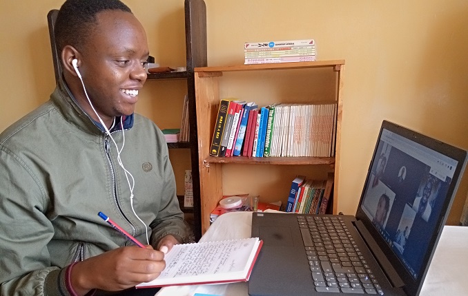 A young man takes notes while engaging in an online meeting through a computer on his desk