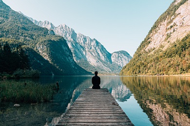 Una persona sentada en un muelle mirando una masa de agua y montañas
