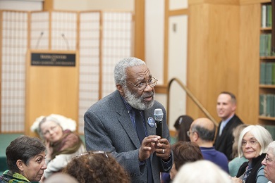 An elderly African American man addressing an audience in a hall.