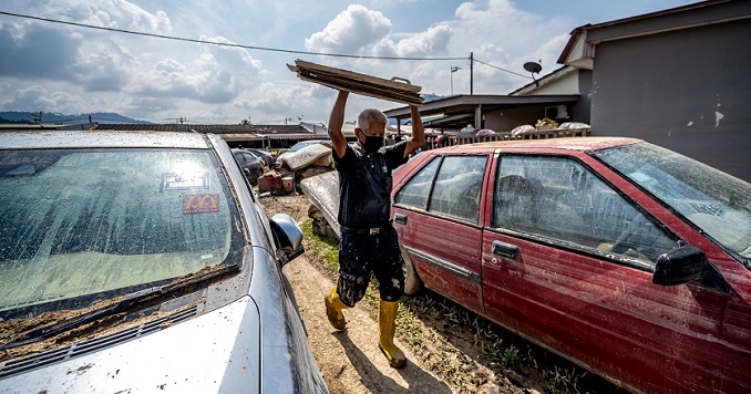 一名男性在头顶上举着几片木板，走在两辆被洪水毁坏的汽车之间。