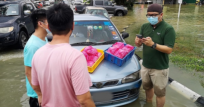 Three people stand ankle-deep in water next to a car in a flooded road.