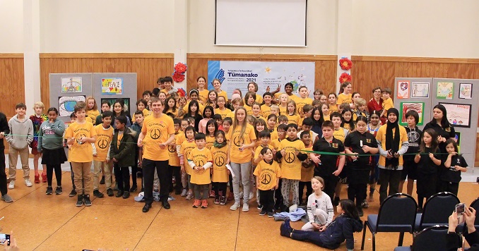 A large group of children and some adults in matching T-shirts pose for a photo in a hall.