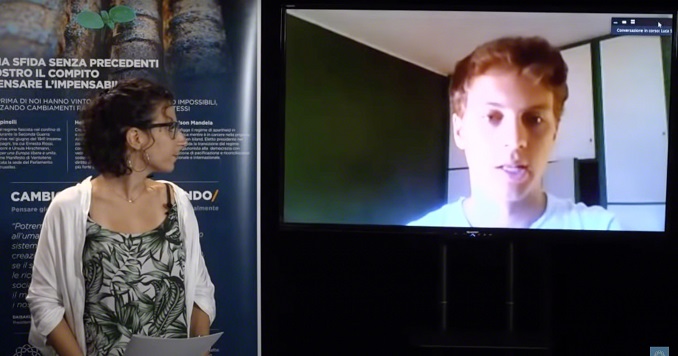 A woman presenting from in front of an exhibition panel looks at a large screen on which a man is speaking