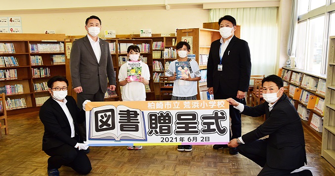 A group of two students and four adults pose behind a small banner written in Japanese
