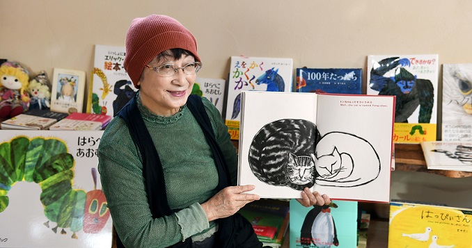 Yoshiko Isoda, seated in front of a shelf of children’s books, holds open a picture book