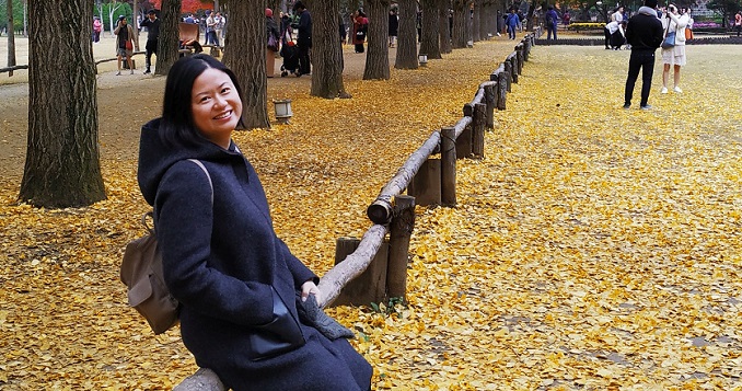 Delante de una hilera de árboles, Yen Gan está sentada en una valla sobre el suelo cubierto de hojas amarillas