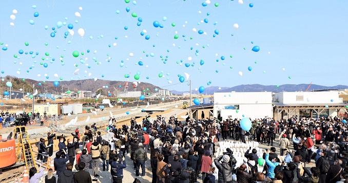 A crowd watches white, blue and green balloons float away